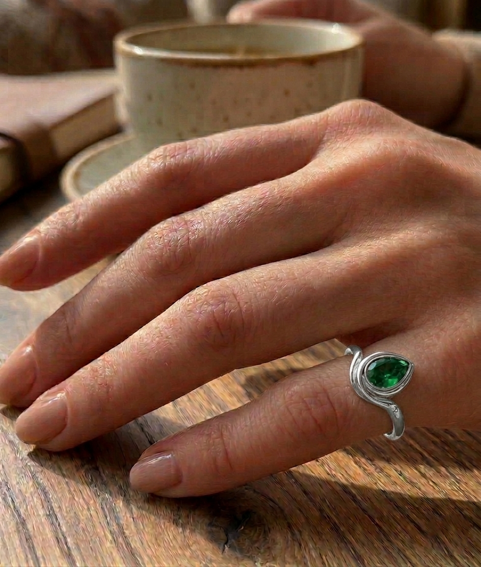 Hand wearing a white gold ring with a green gemstone on a wooden surface with a cup in the background.