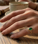 Hand wearing a gold ring with a green gemstone on a wooden surface with a cup in the background