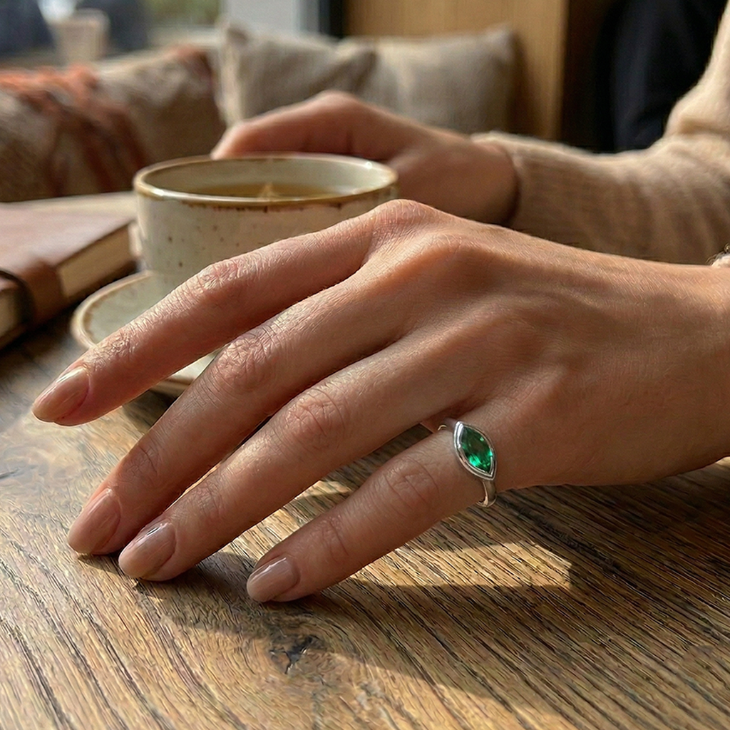 Hand with a green gemstone ring on a wooden table with a cup in the background