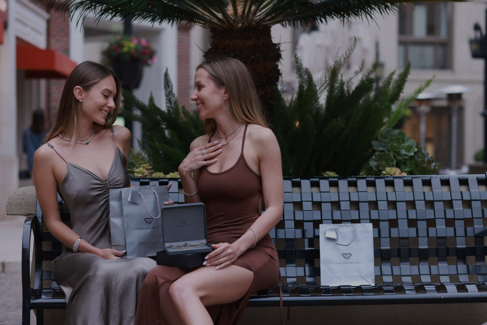 Two women sitting on a bench with a box and bags, possibly at an outdoor shopping area.